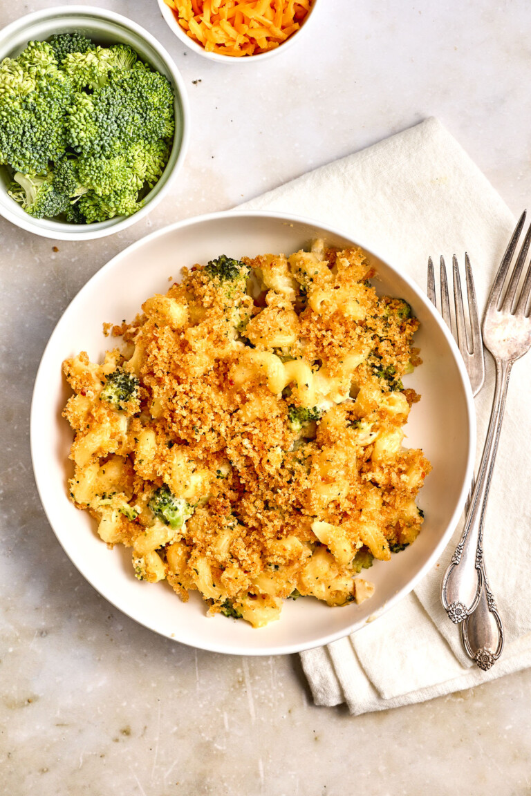 A bowl of broccoli macaroni and cheese topped with breadcrumbs, placed on a napkin with two forks, with a bowl of raw broccoli and a bowl of shredded cheese nearby.
