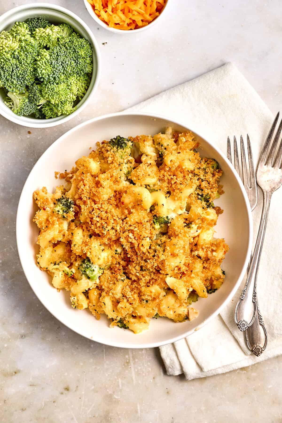 A bowl of broccoli macaroni and cheese topped with breadcrumbs, placed on a napkin with two forks, with a bowl of raw broccoli and a bowl of shredded cheese nearby.