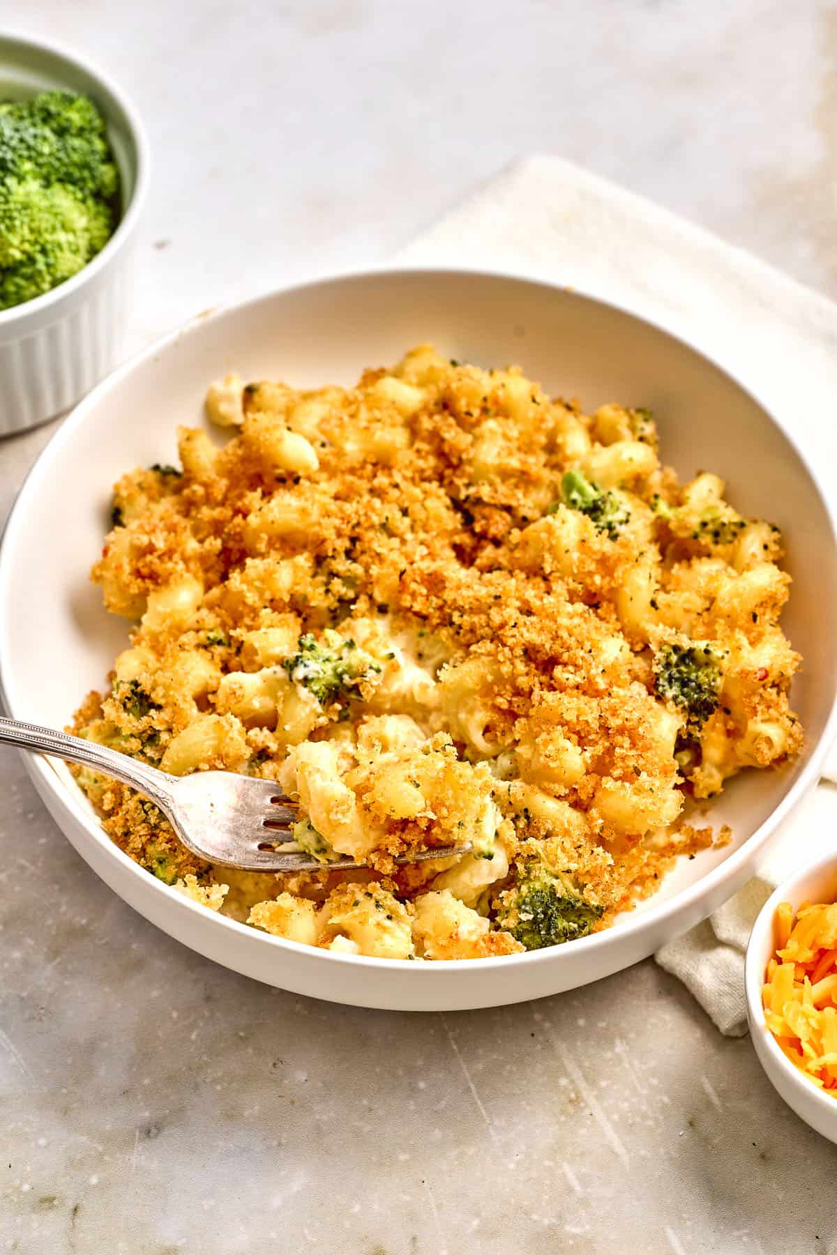Bowl of baked macaroni and cheese with broccoli, topped with golden breadcrumbs, with a fork in the dish. Small bowls of broccoli and shredded cheese are nearby.