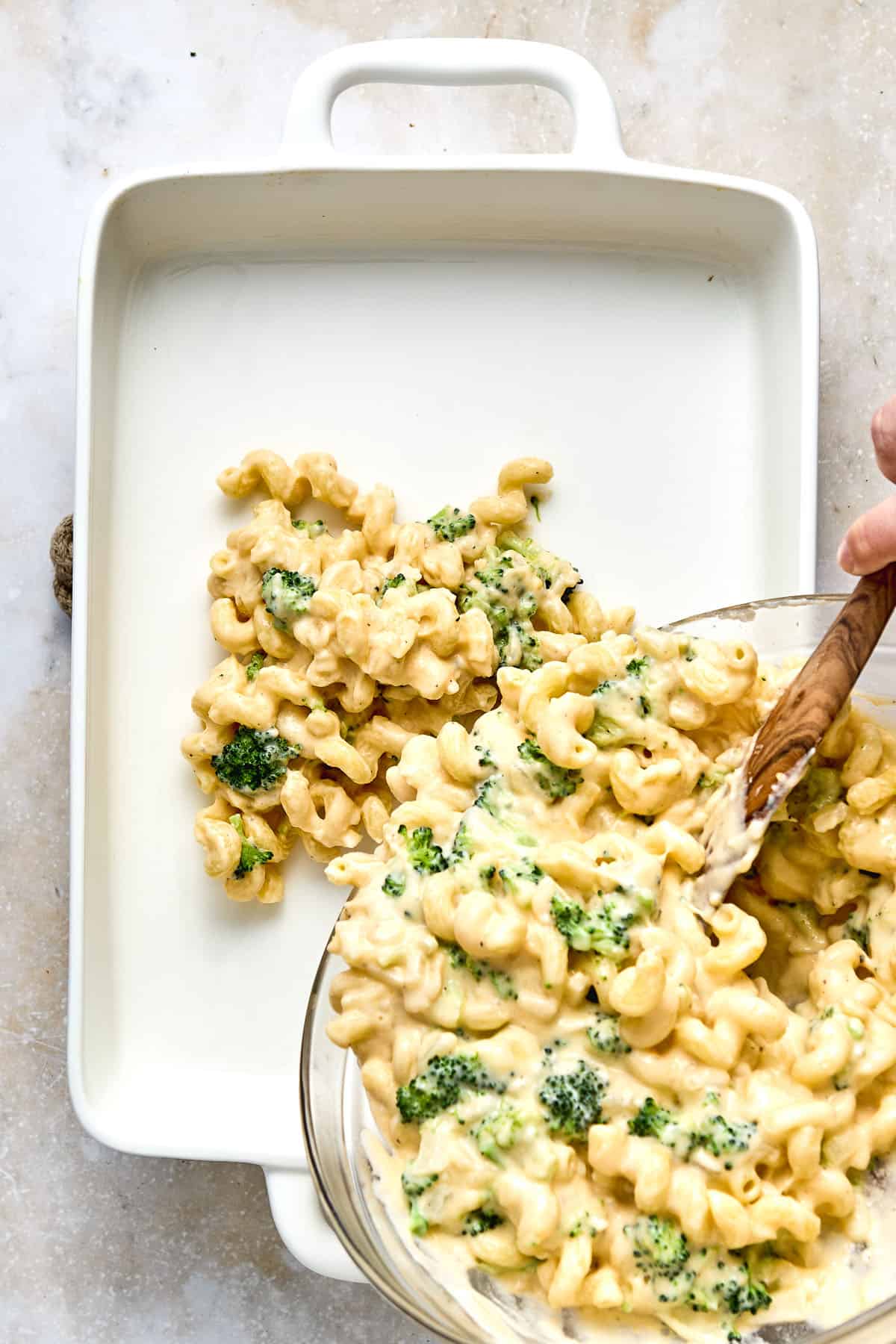 A hand pours broccoli macaroni and cheese from a bowl into a white rectangular baking dish on a marble surface.