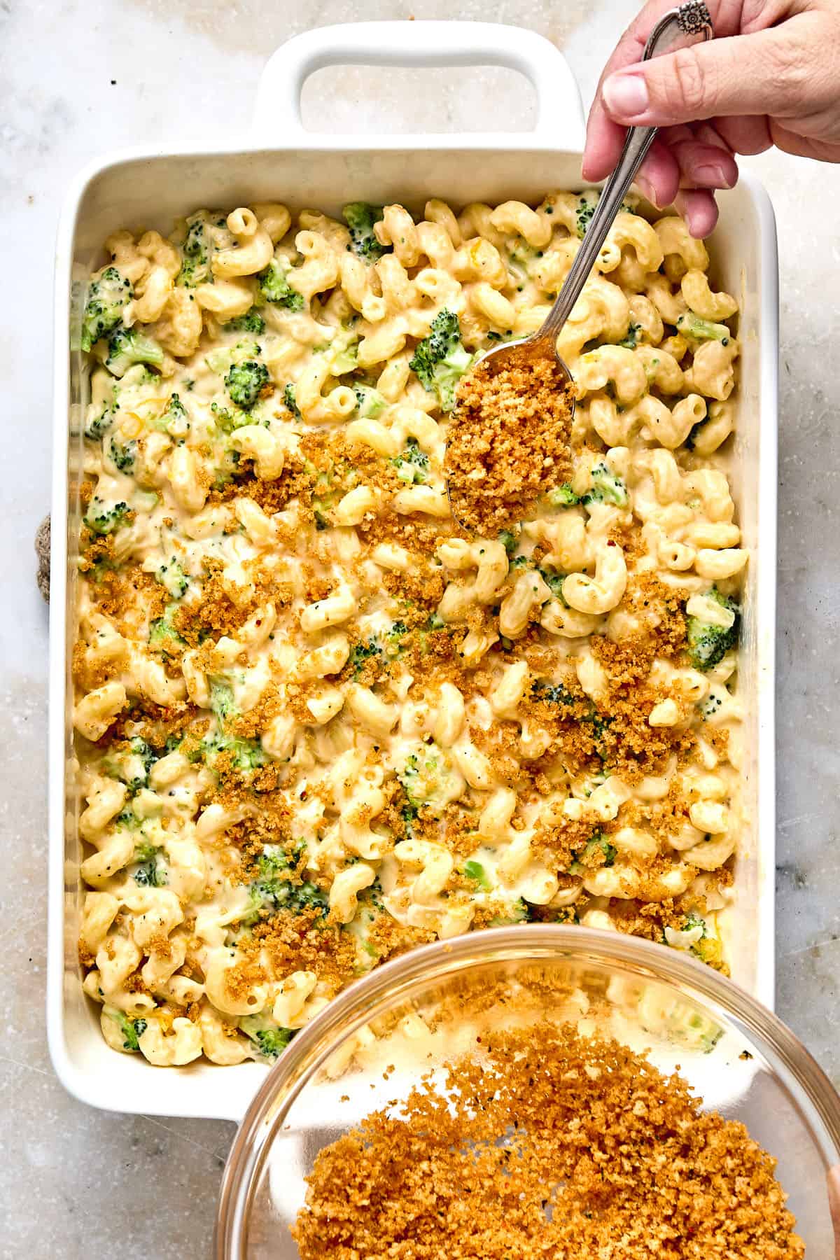 A hand sprinkles breadcrumb topping over a casserole dish filled with macaroni, cheese sauce, and broccoli.