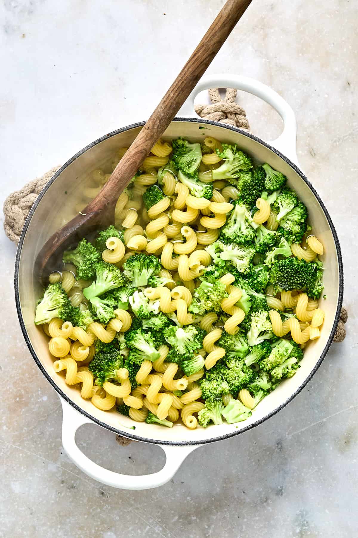 A white pot filled with cooked pasta and broccoli, with a wooden spoon resting inside, on a light-colored surface.