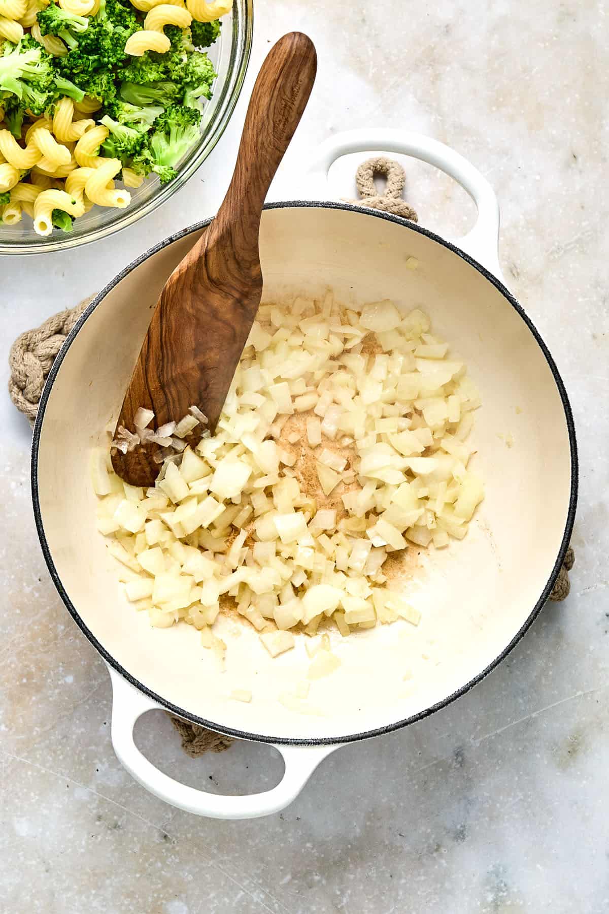 Chopped onions cooking in a white pot with a wooden spoon; a bowl of pasta and broccoli is visible in the background.