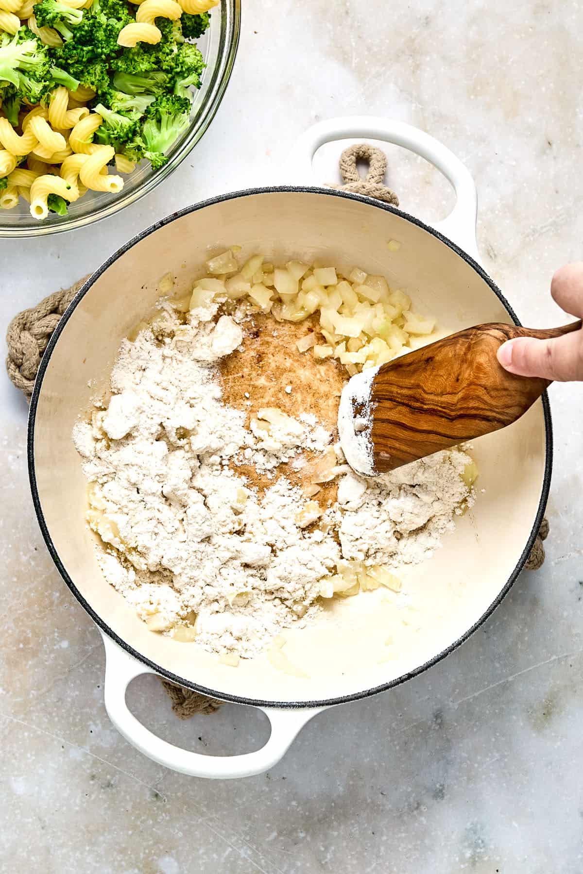 A hand stirs chopped onions and flour in a white pot with a wooden spatula; a bowl of pasta and broccoli is visible nearby.
