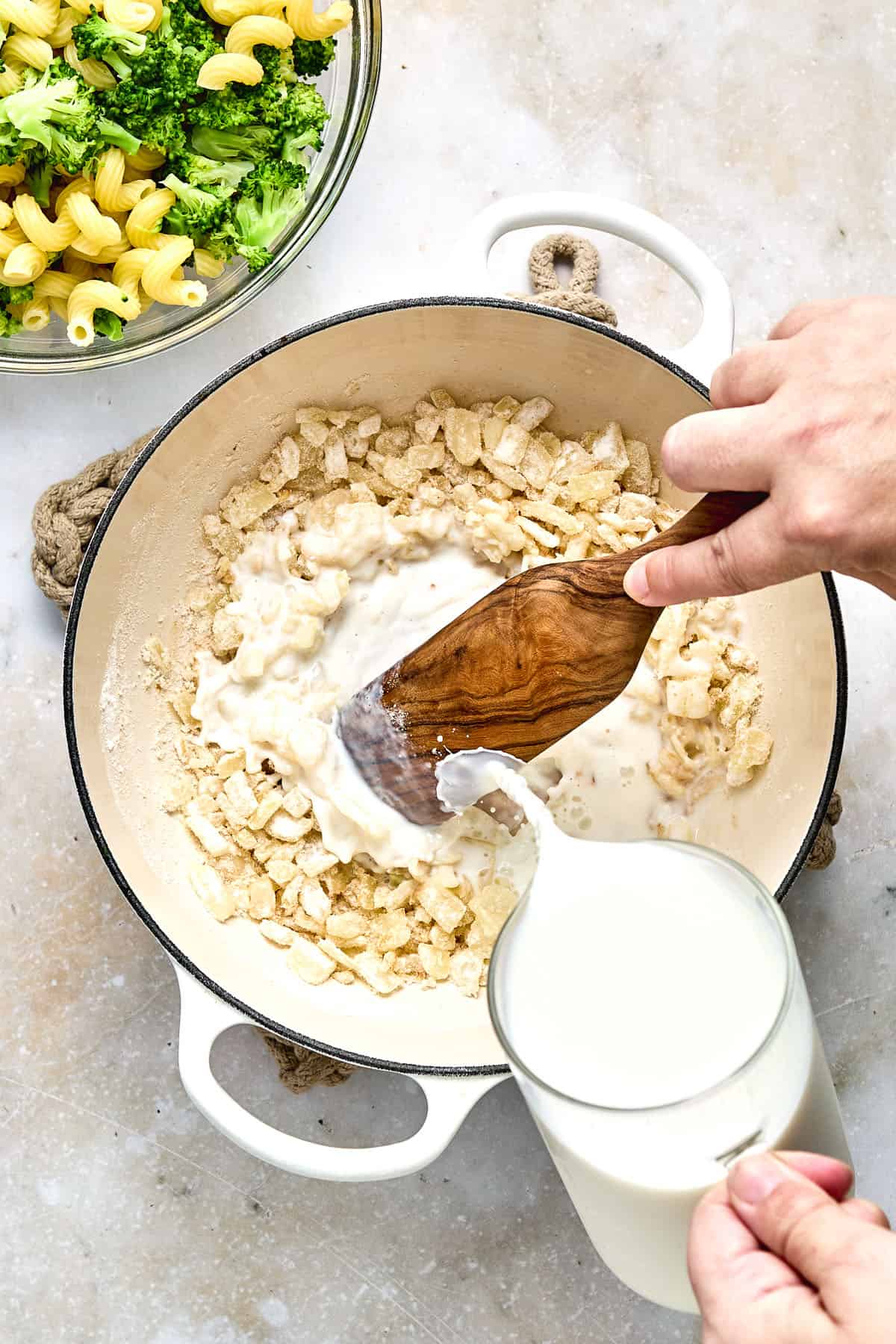 A person stirs flour and butter in a white pot while pouring in milk; cooked pasta and broccoli are in a bowl nearby.