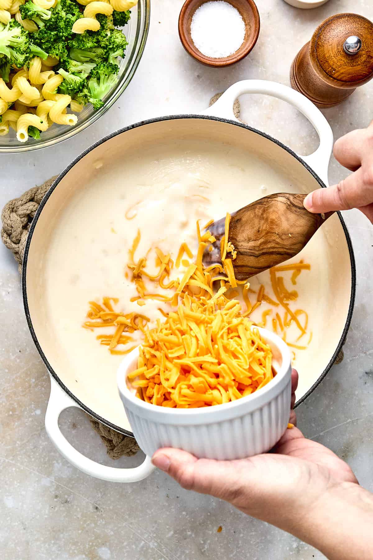 A person adds shredded cheddar cheese from a white bowl into a saucepan of creamy sauce, with cooked pasta and broccoli visible nearby.