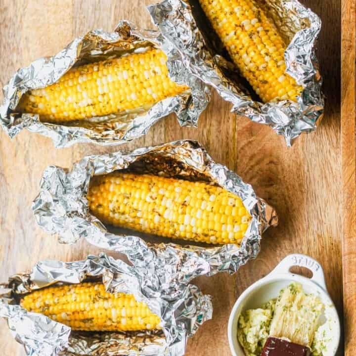 Four ears of corn on the cob wrapped in aluminum foil on a wooden surface, with a small dish of butter and seasonings nearby.