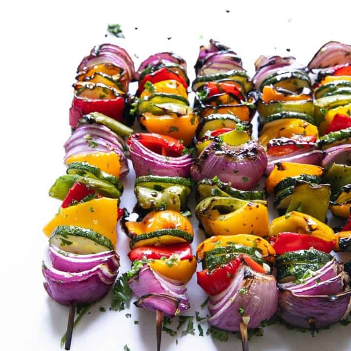 Grilled vegetable skewers with red onion, bell peppers, and zucchini on wooden sticks, garnished with chopped herbs, arranged on a white background.