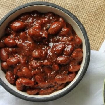 A bowl filled with baked beans in a thick, reddish-brown sauce, placed on a white surface with part of a burlap fabric visible.