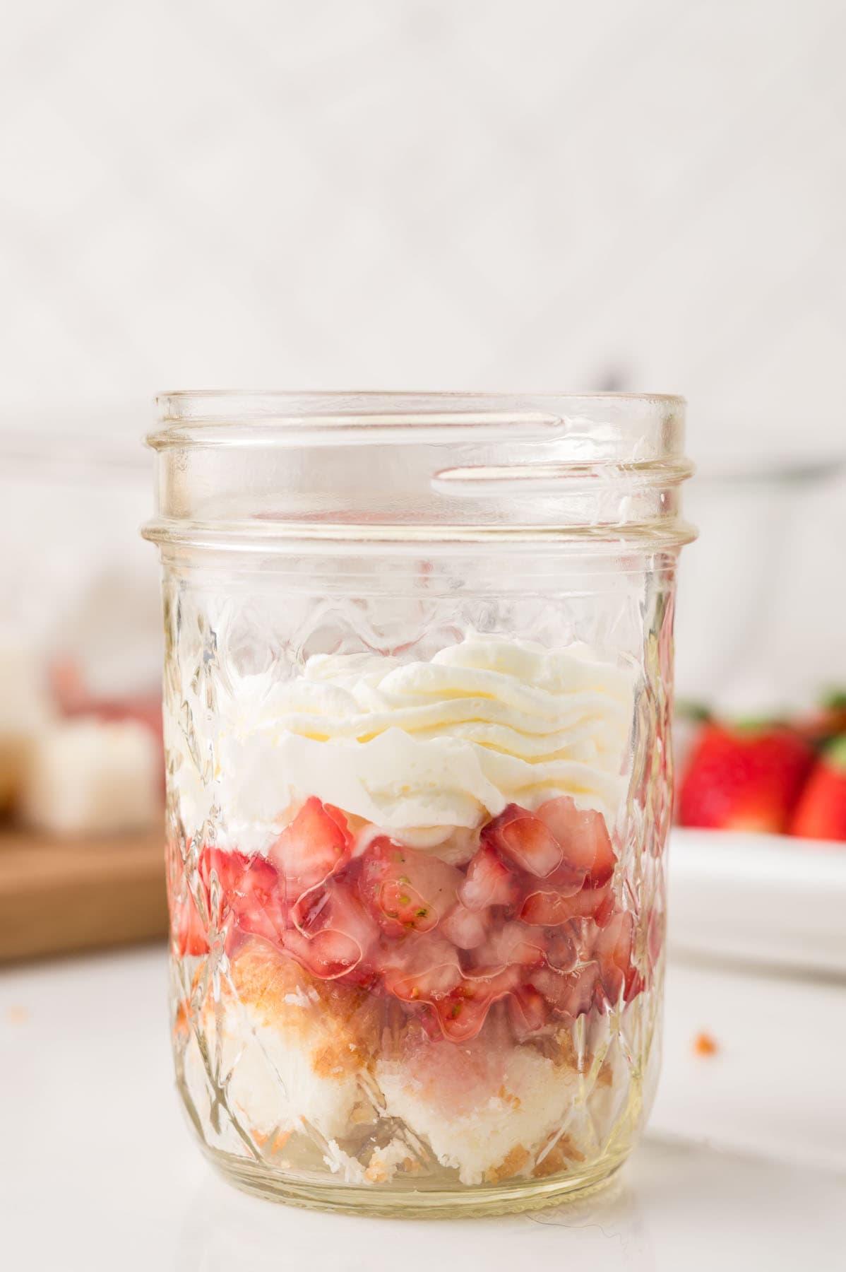A glass jar filled with layers of cubed cake, chopped strawberries, and whipped cream sits on a white surface.