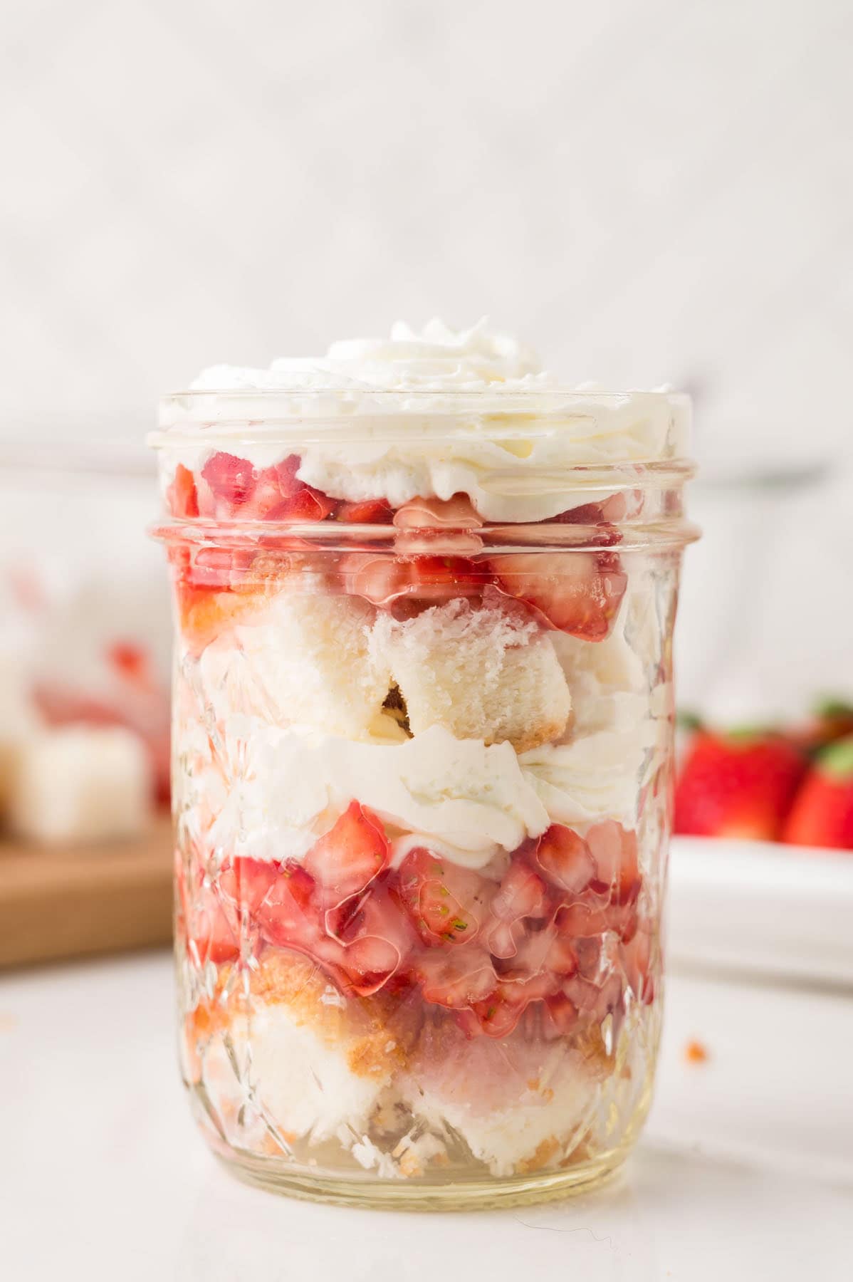A mason jar filled with layers of whipped cream, chopped strawberries, and pieces of cake, with more strawberries visible in the background.
