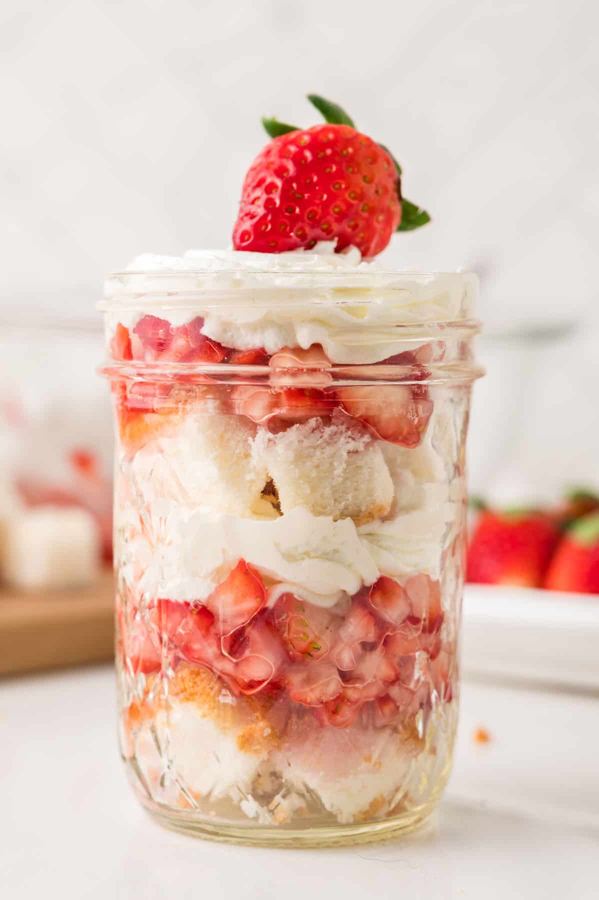 A mason jar filled with layers of whipped cream, chopped strawberries, and cake pieces, topped with whipped cream and a whole strawberry.