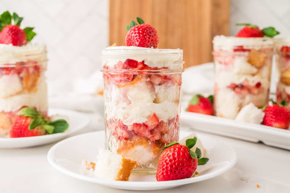 Individual dessert in a glass jar filled with layers of whipped cream, diced strawberries, and cake pieces, topped with a whole strawberry.