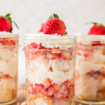 Three glass jars filled with layered strawberry shortcake, whipped cream, and sponge cake, each topped with a whole strawberry, are placed on a wooden surface.