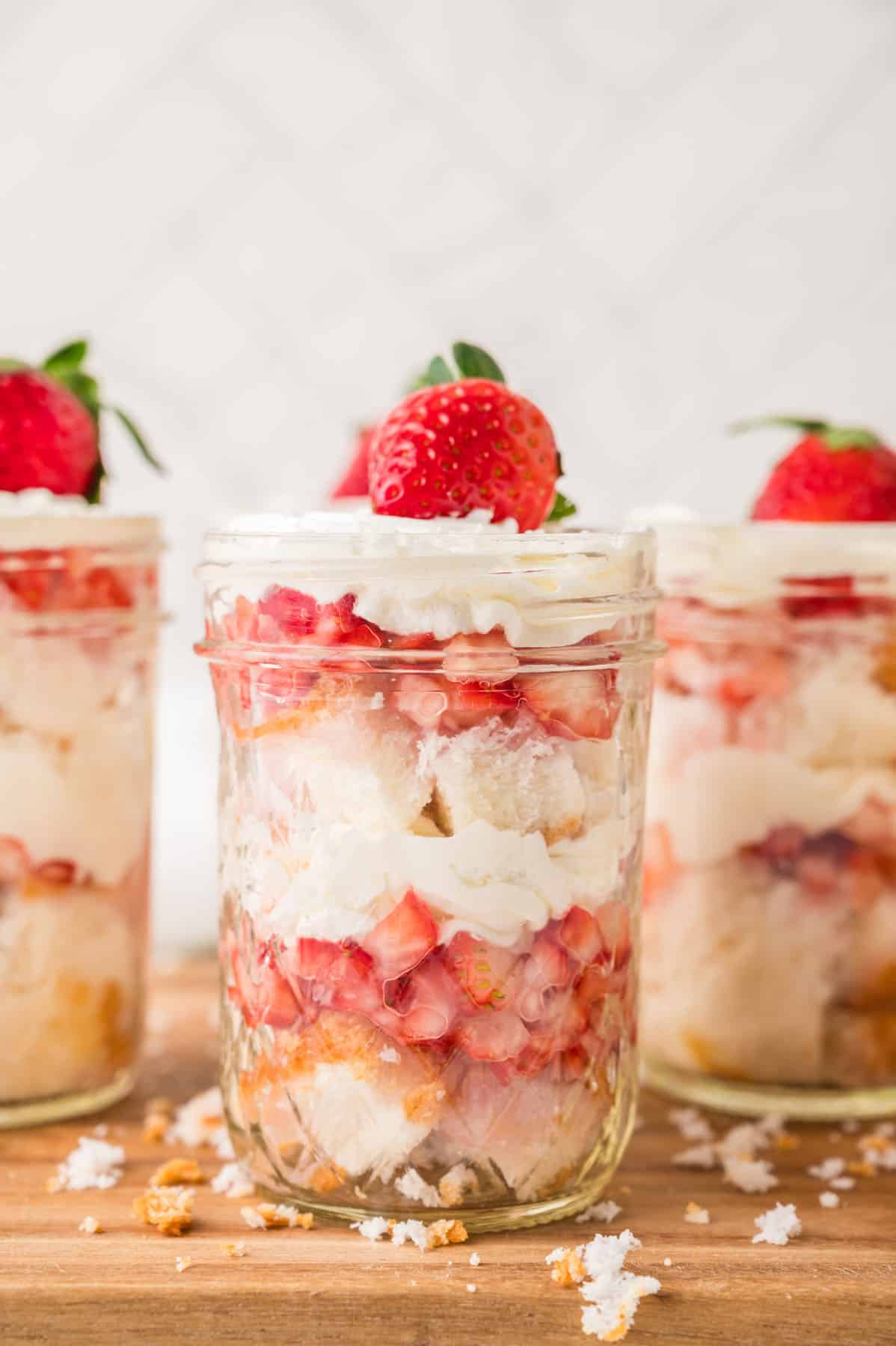 Three glass jars filled with layered strawberry shortcake, whipped cream, and sponge cake, each topped with a whole strawberry, are placed on a wooden surface.