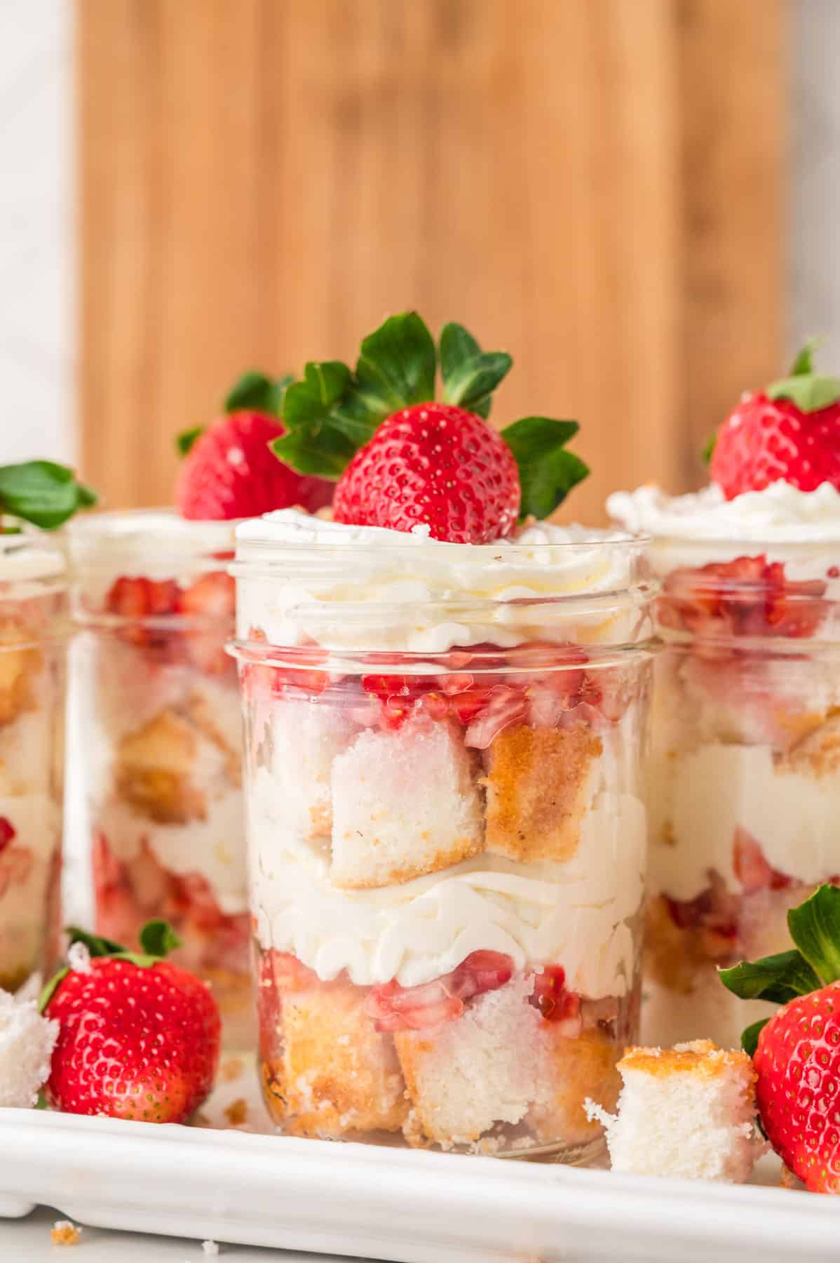 Individual glass jars filled with layers of cubed cake, whipped cream, and strawberries, topped with a whole fresh strawberry, arranged on a tray.