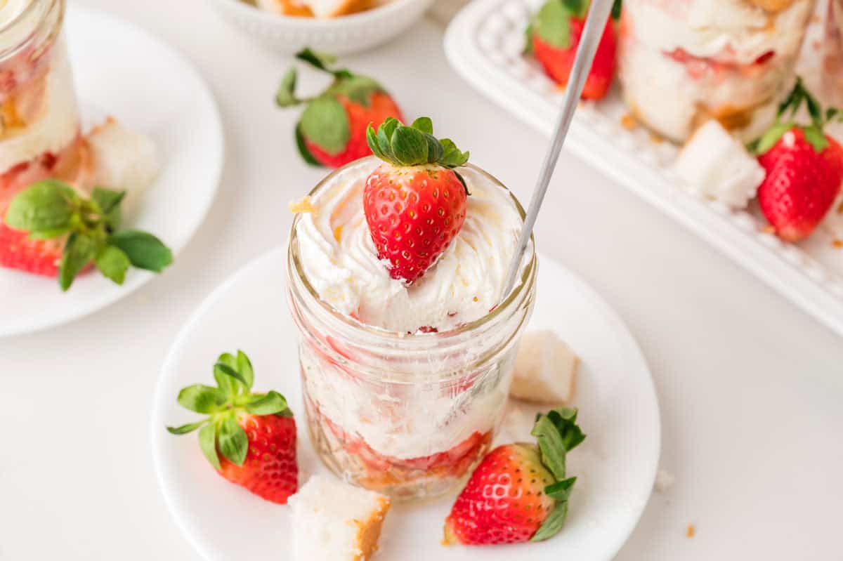 A glass jar filled with whipped cream, cake pieces, and strawberries, topped with a whole strawberry, is placed on a white plate with more strawberries and cake pieces around it.