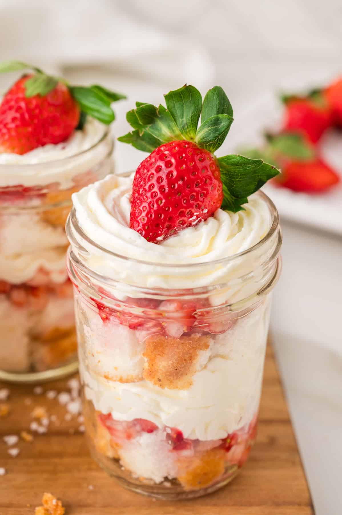Layered strawberry shortcake dessert jars with layers of whipped cream, diced macerated strawberries, angel food cake pieces, and topped with a whole strawberry.