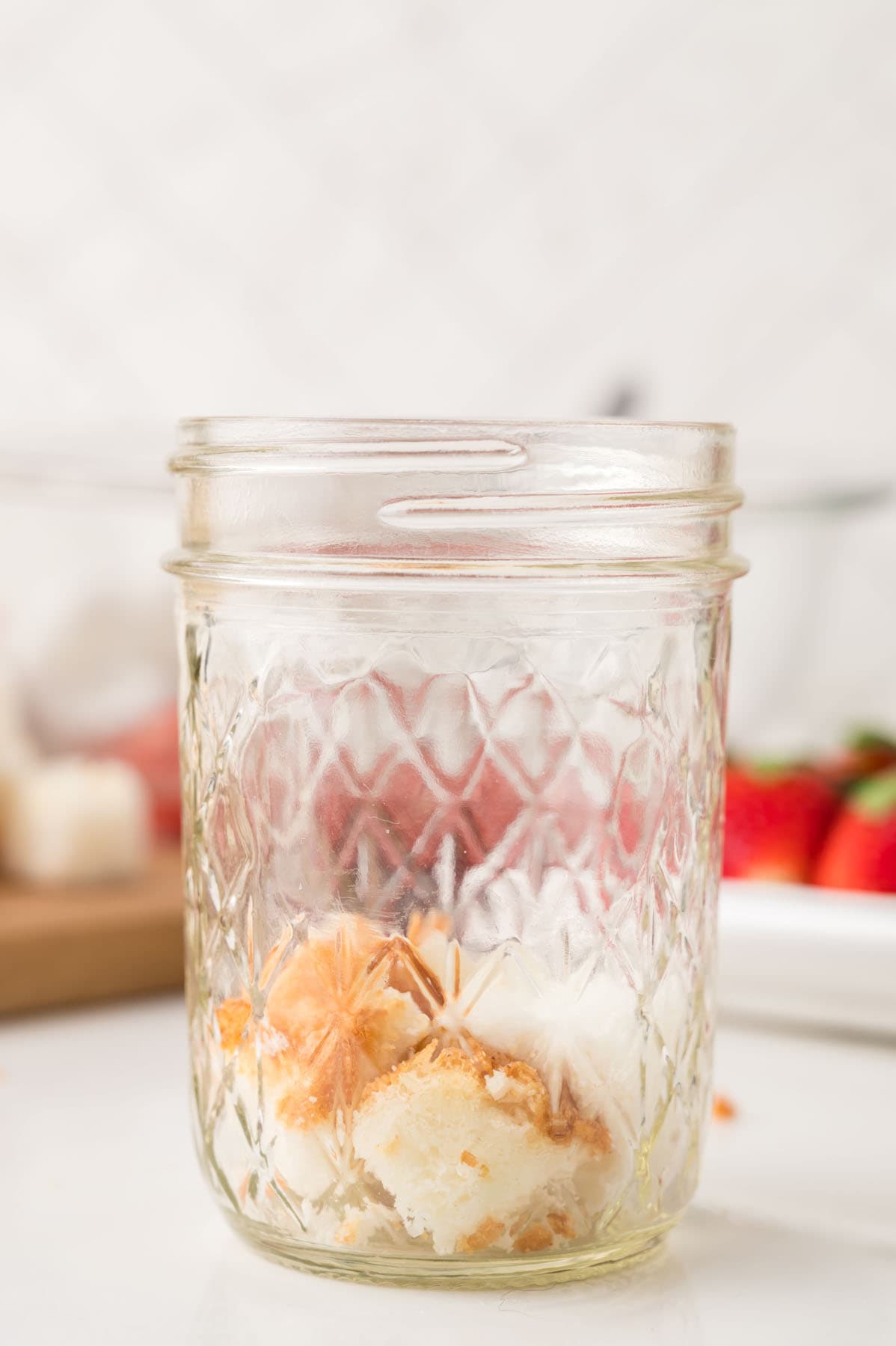 A glass mason jar containing pieces of yellow and white cake, with strawberries and a cutting board in the blurred background.