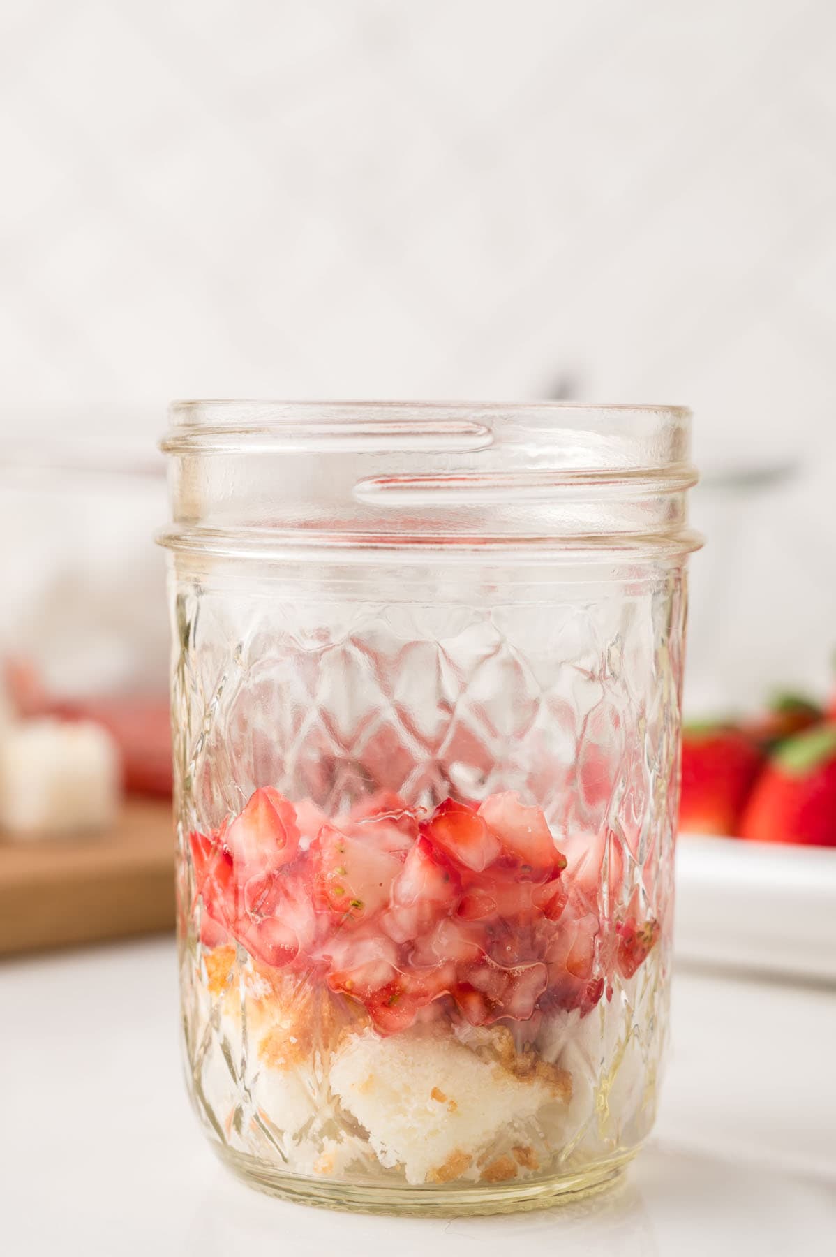 A mason jar filled with chopped strawberries and pieces of cake sits on a white surface, with whole strawberries and a cutting board in the background.