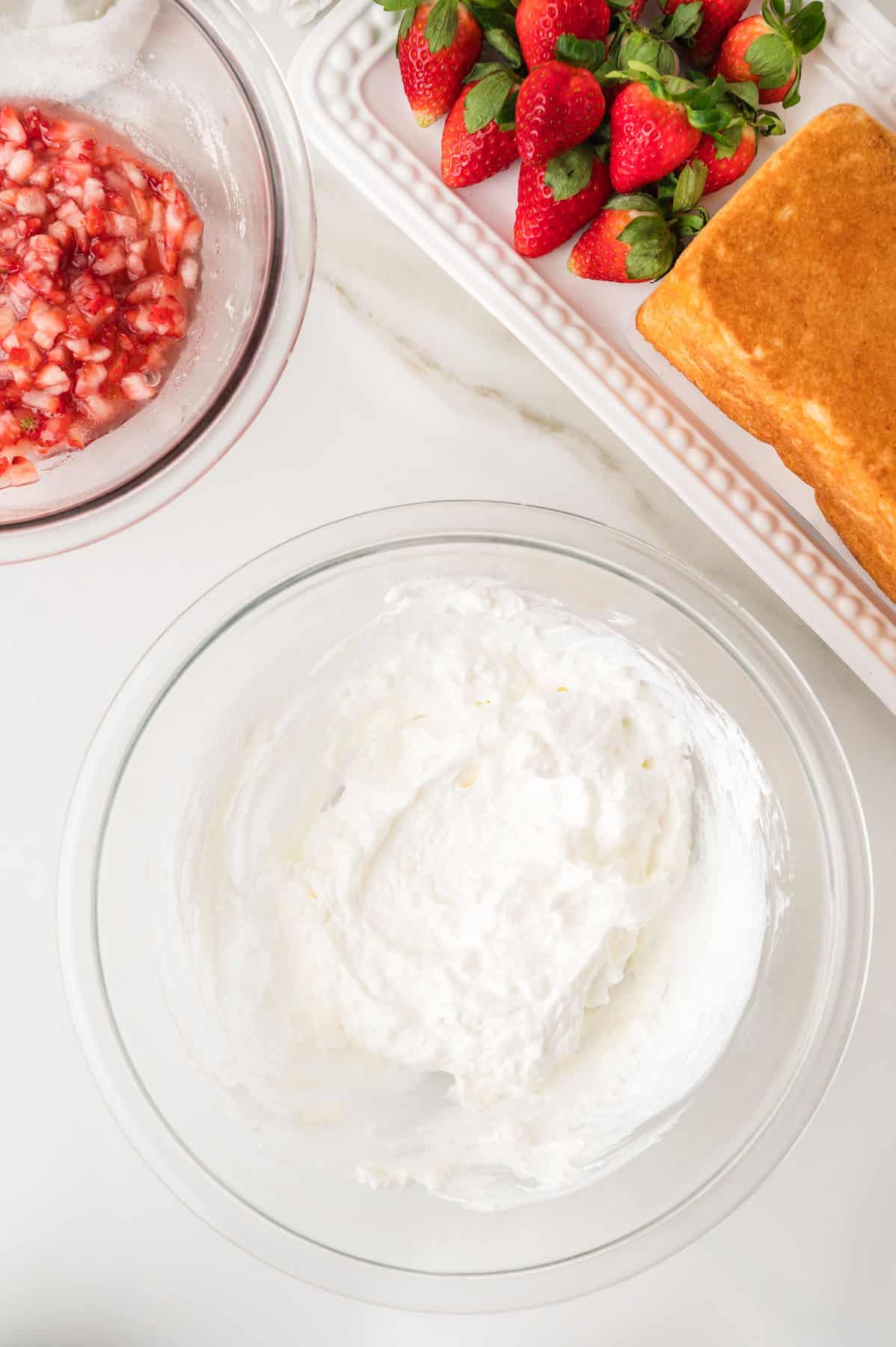 A bowl of whipped cream, a bowl of chopped strawberries, whole strawberries, and slices of pound cake arranged on a white surface.
