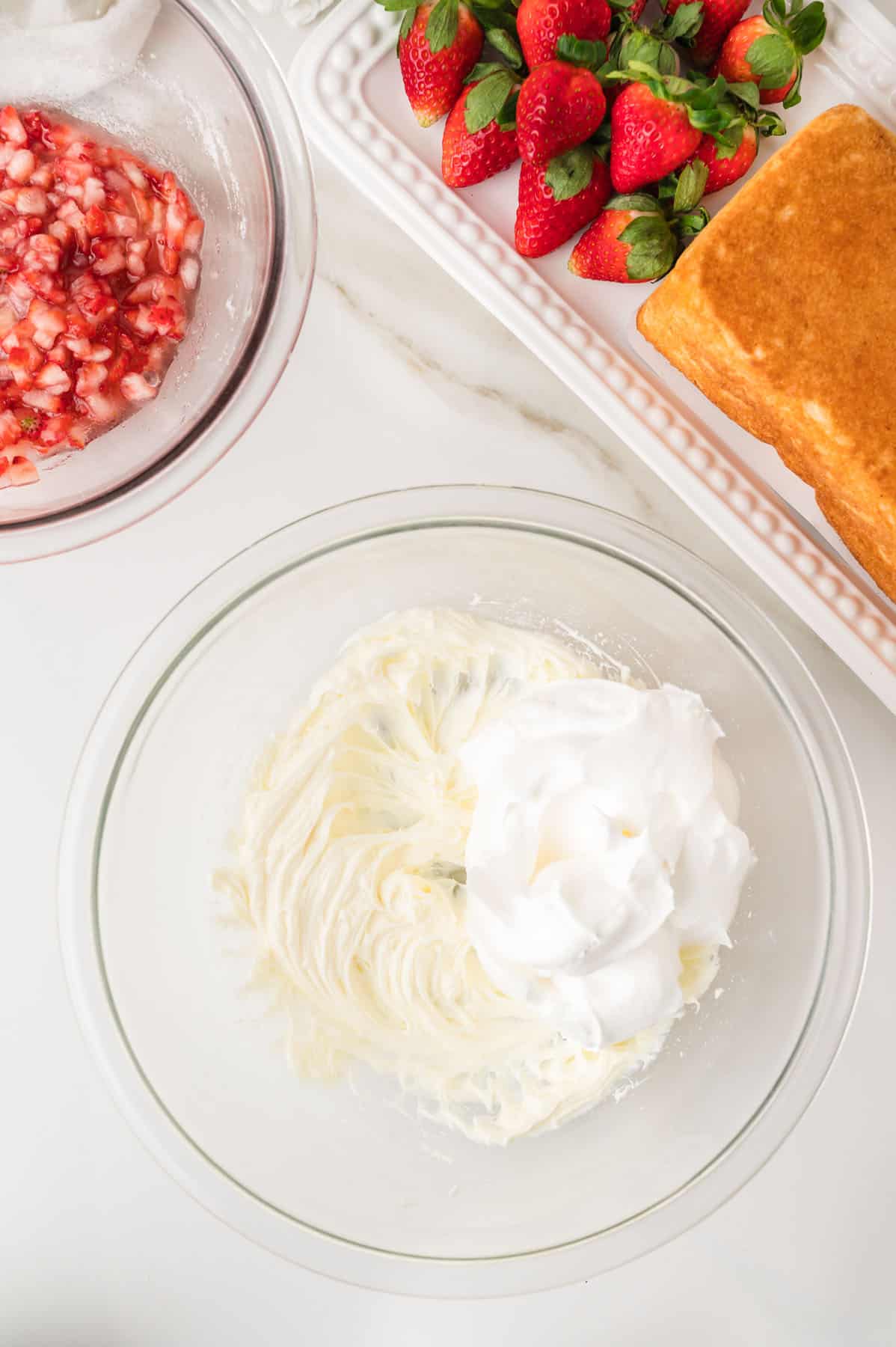 A glass bowl with whipped cream and cream cheese mixture, a bowl of chopped strawberries, and a tray with whole strawberries and sliced pound cake on a white surface.