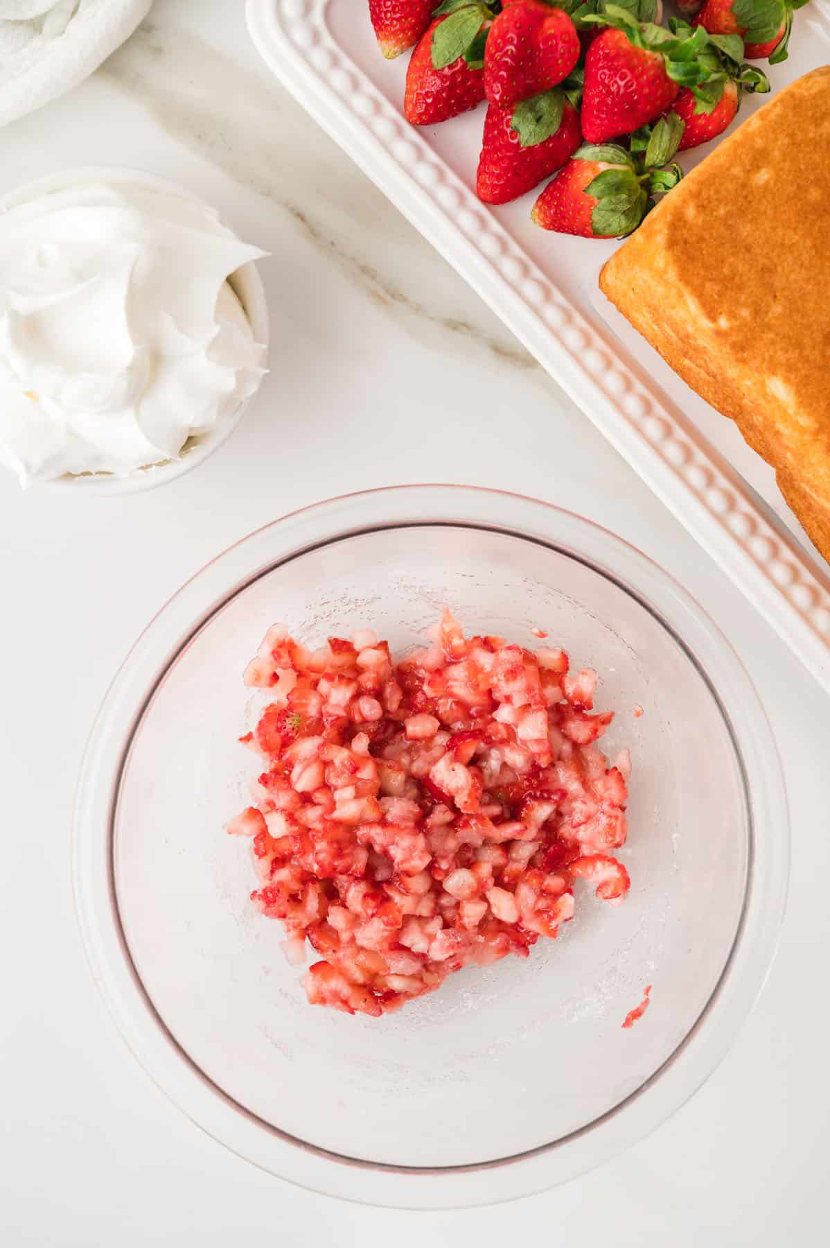 Chopped macerated strawberries in a glass bowl, with whole strawberries, a loaf of cake, and whipped cream nearby on a white surface.