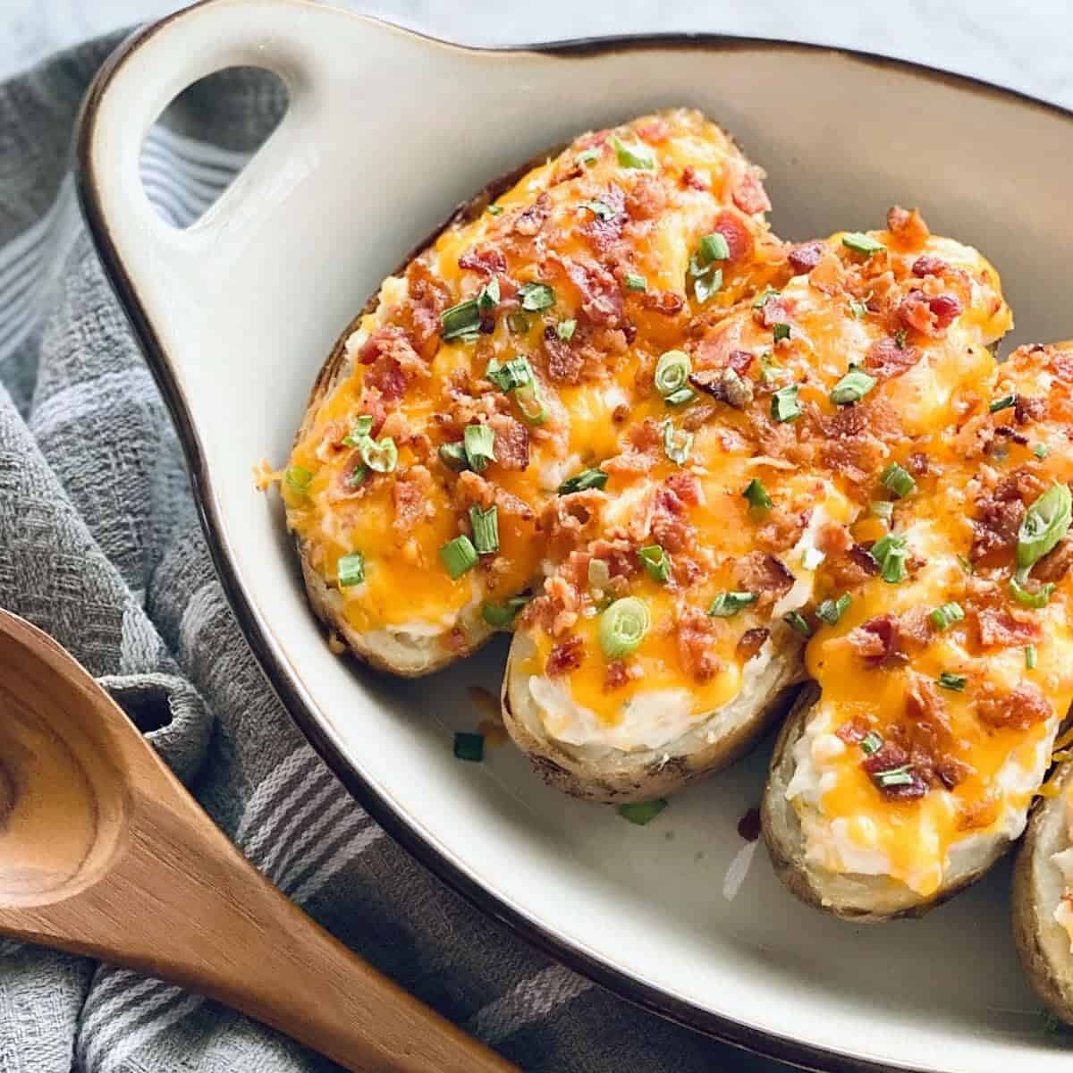 Three twice-baked potatoes topped with melted cheese, bacon bits, and chopped green onions in a white baking dish next to a wooden spoon.