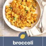 A bowl of broccoli mac and cheese topped with golden breadcrumbs sits on a marble surface beside a fork, spoon, and white napkin.