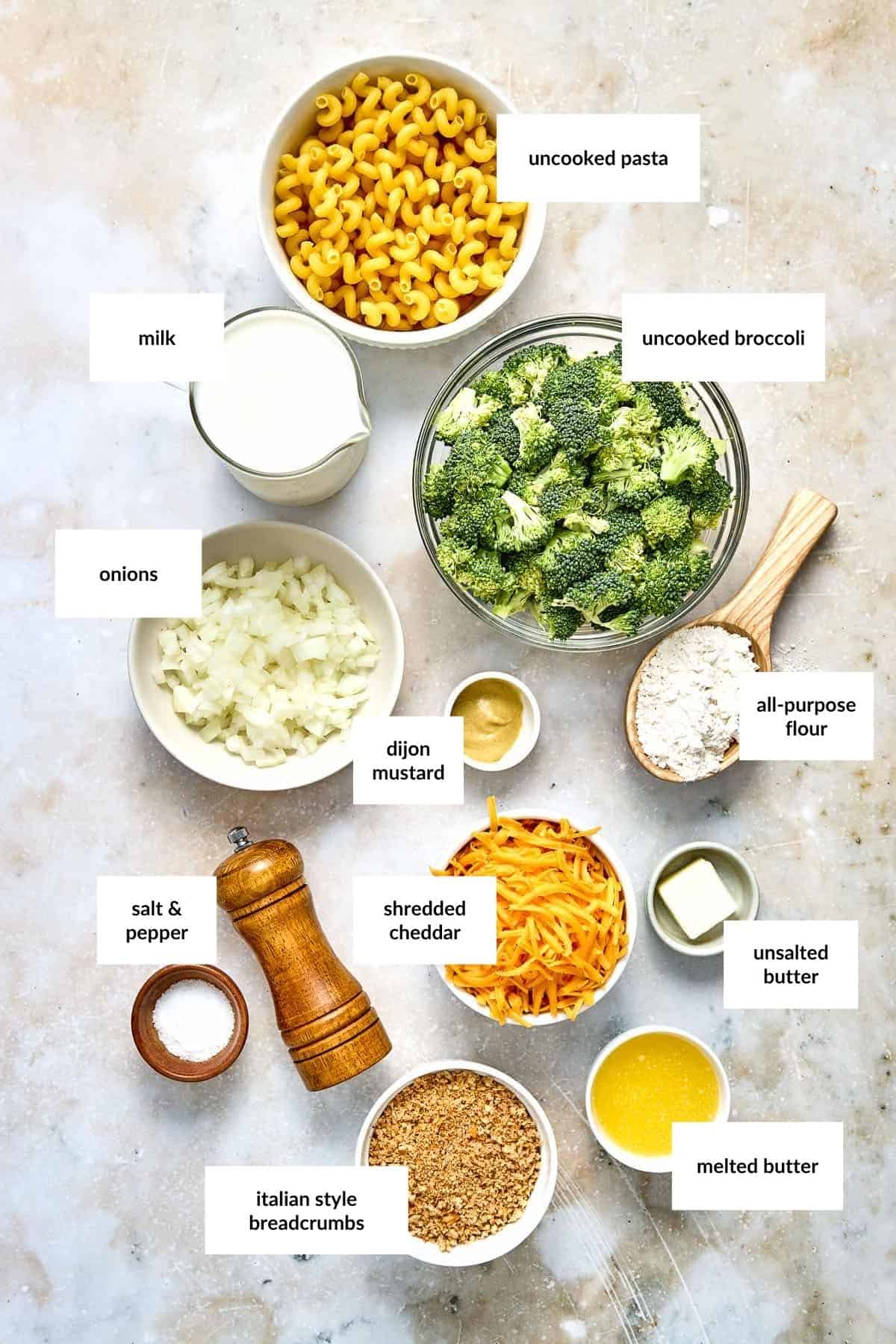 An overhead view of labeled ingredients for a broccoli cheddar pasta bake, including uncooked pasta, broccoli, milk, onions, dijon mustard, flour, cheddar, butter, breadcrumbs, salt, and pepper.