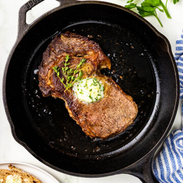 A cooked steak topped with herb butter sits in a cast iron skillet, with fresh parsley and buttered bread on the side.