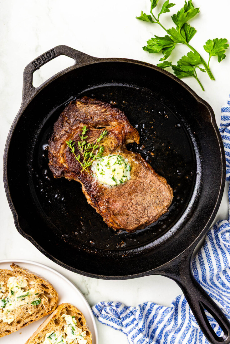 A cooked steak topped with herb butter sits in a cast iron skillet, with fresh parsley and buttered bread on the side.