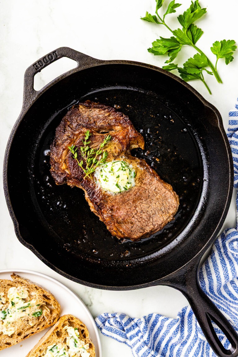 A cooked steak topped with herb butter sits in a cast iron skillet, with fresh parsley and buttered bread on the side.