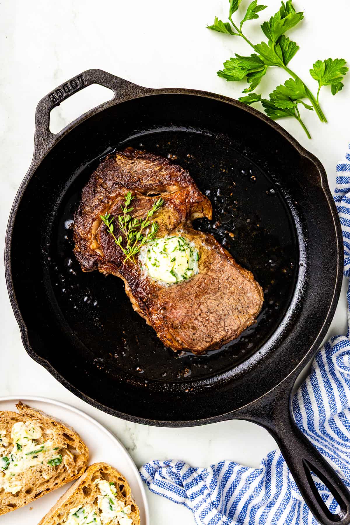 A cooked steak topped with herb butter sits in a cast iron skillet, with fresh parsley and buttered bread on the side.