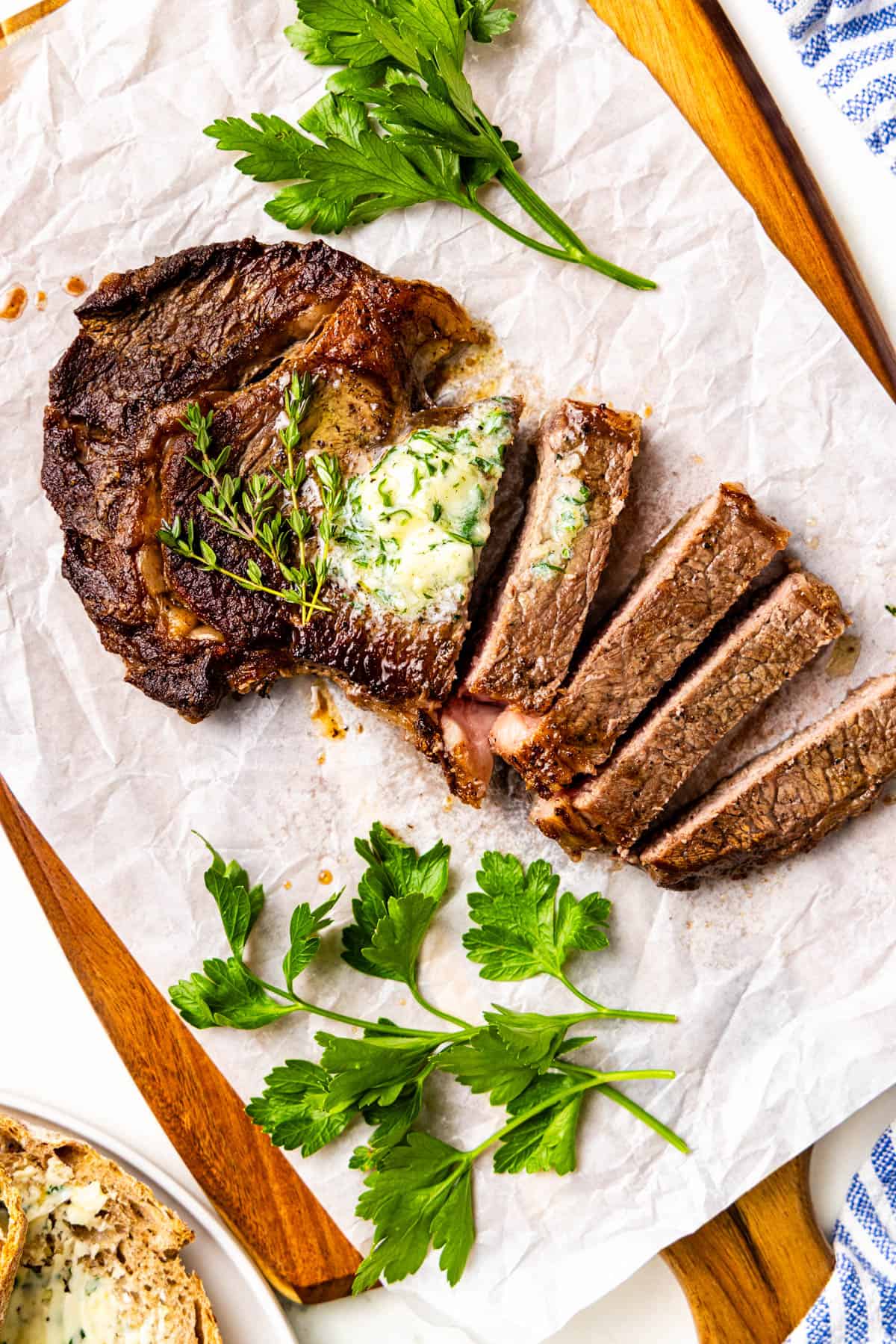 A cooked steak topped with herb butter is partially sliced and garnished with fresh parsley on parchment paper over a wooden board.