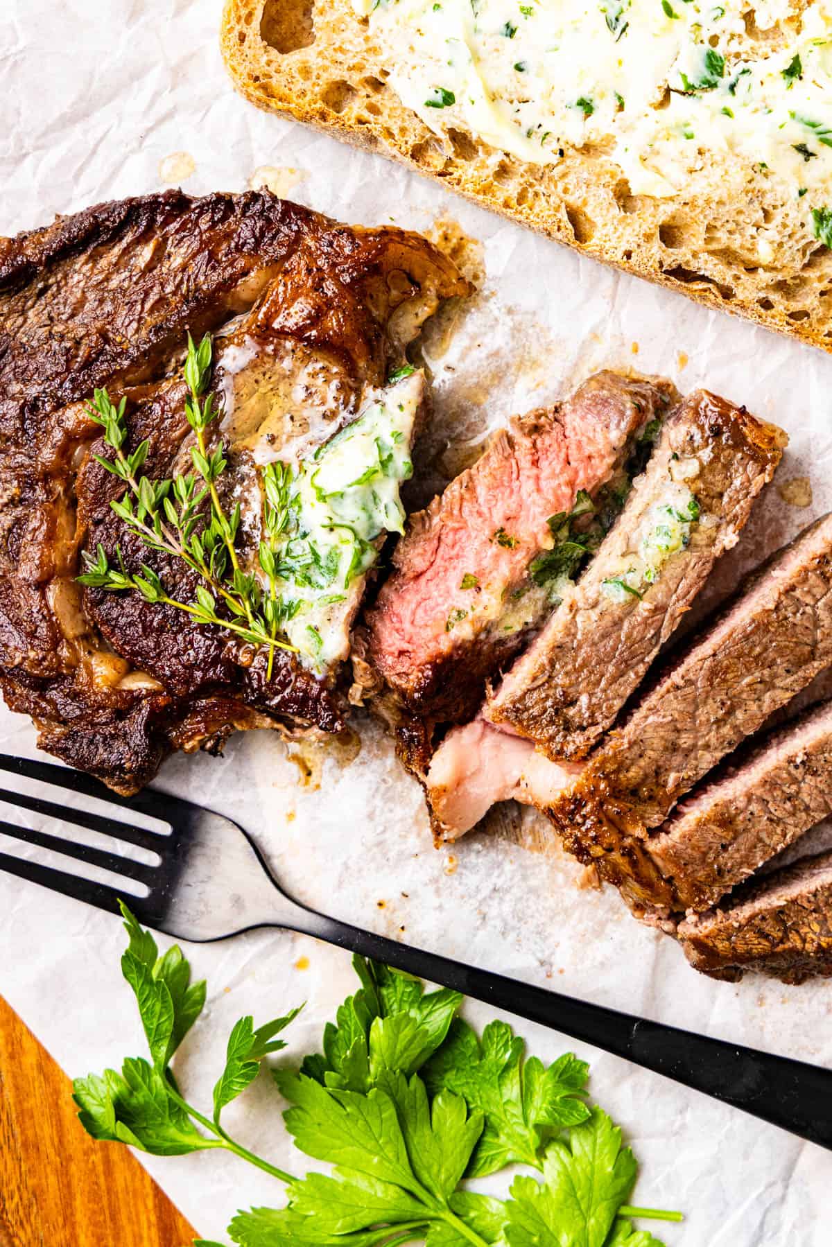 Sliced cooked steak topped with herb butter, garnished with fresh herbs, next to a slice of bread with herb butter and a fork on parchment paper.