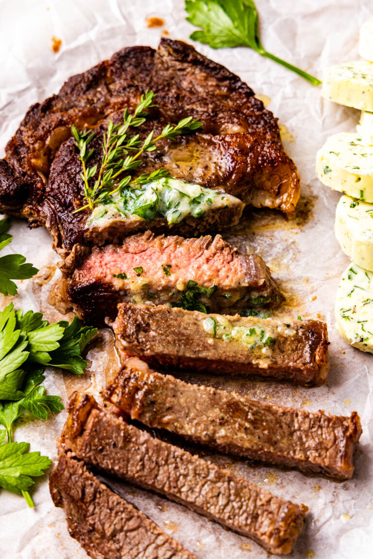 Sliced cooked steak topped with herb butter and garnished with fresh herbs, displayed on parchment paper with extra herb butter and parsley on the side.
