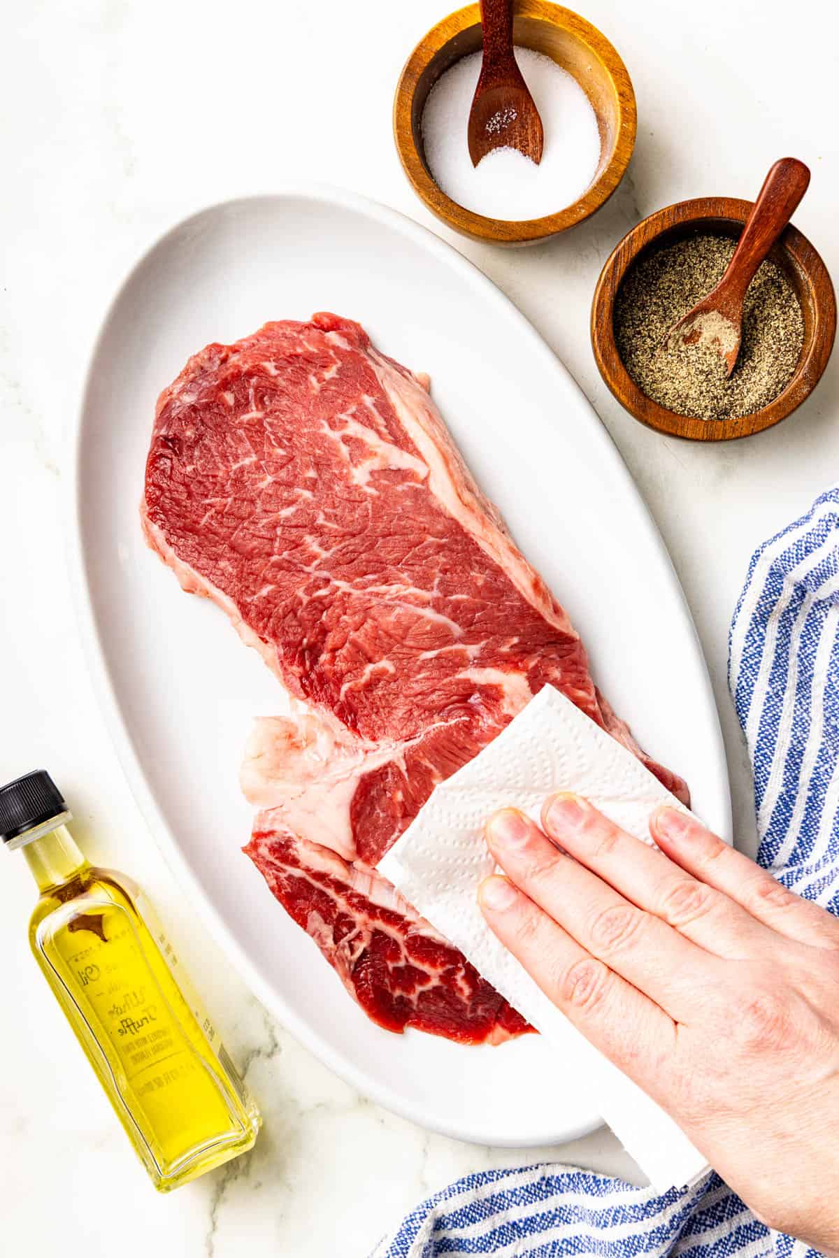 A hand pats raw steak with a paper towel on a white plate, next to bowls of salt, pepper, and a bottle of olive oil.