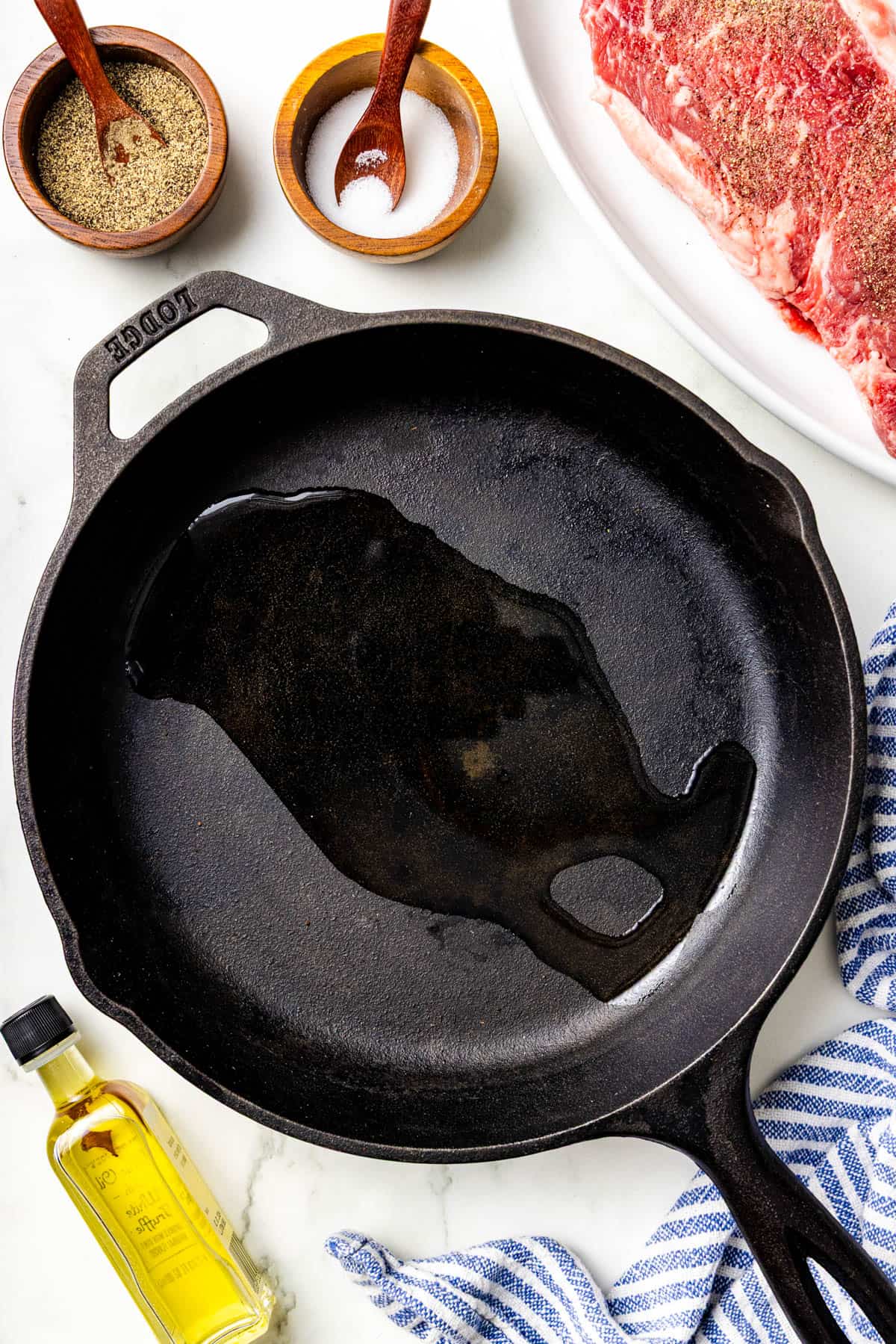 A cast iron skillet with oil sits on a counter near a raw seasoned steak, spices, a blue striped towel, and a bottle of olive oil.