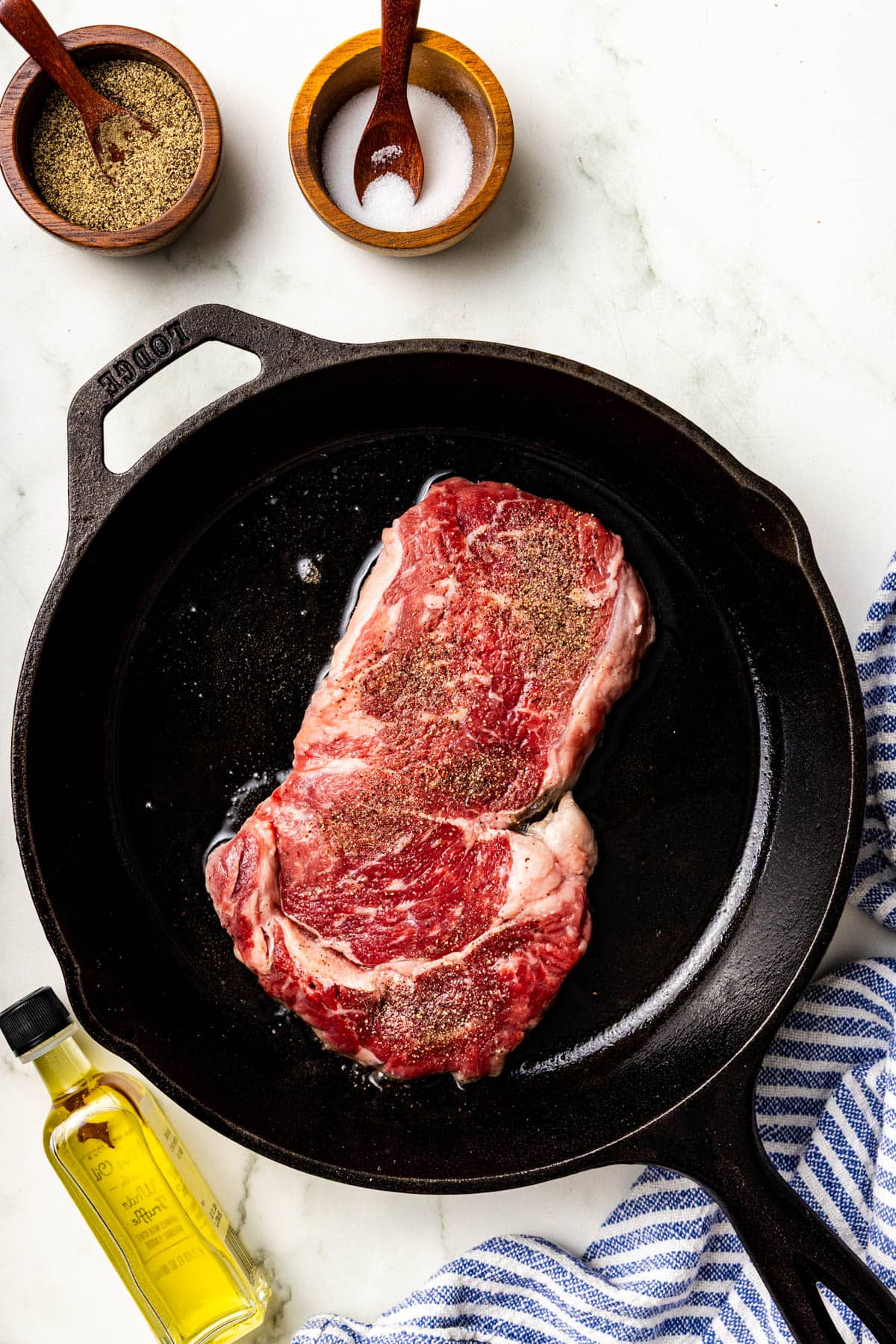 Two raw, seasoned steaks in a cast iron skillet with a bottle of olive oil, a blue striped towel, and small bowls of pepper and salt nearby on a marble surface.
