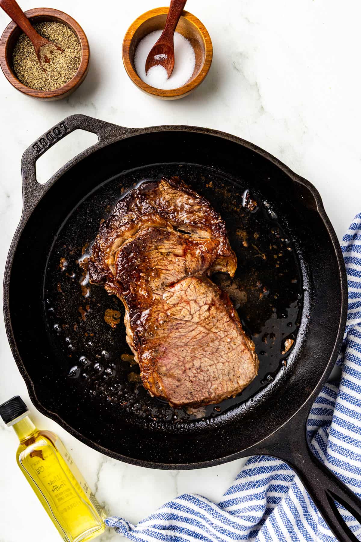 A cooked steak in a cast iron skillet with a bottle of olive oil, bowls of salt and pepper, and a blue-striped towel on a white surface.