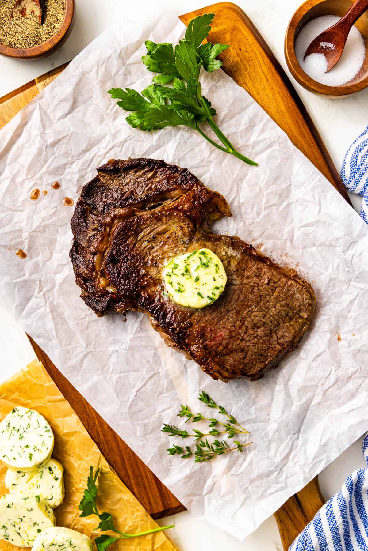Grilled steak topped with a slice of herb butter, garnished with parsley and thyme, served on parchment paper on a wooden board.