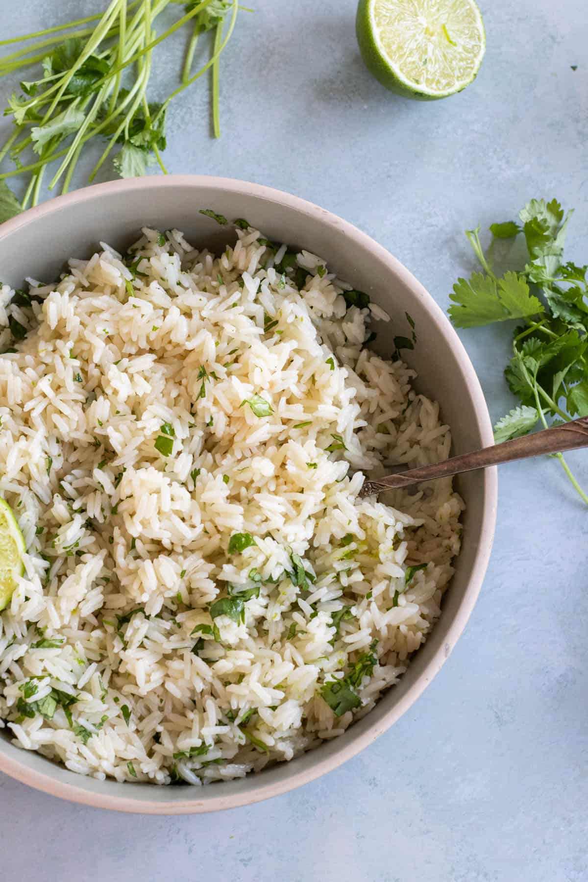 A bowl of white rice mixed with chopped cilantro, with a spoon inside. Fresh cilantro and a halved lime are on the table nearby.