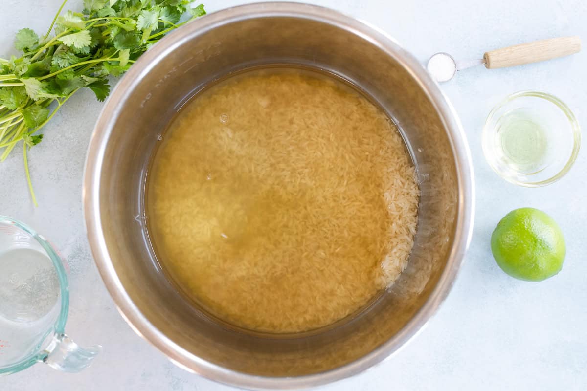 Uncooked rice and water in a metal pot, with cilantro, lime, a measuring cup of water, salt, and a small bowl of oil arranged nearby on a light surface.