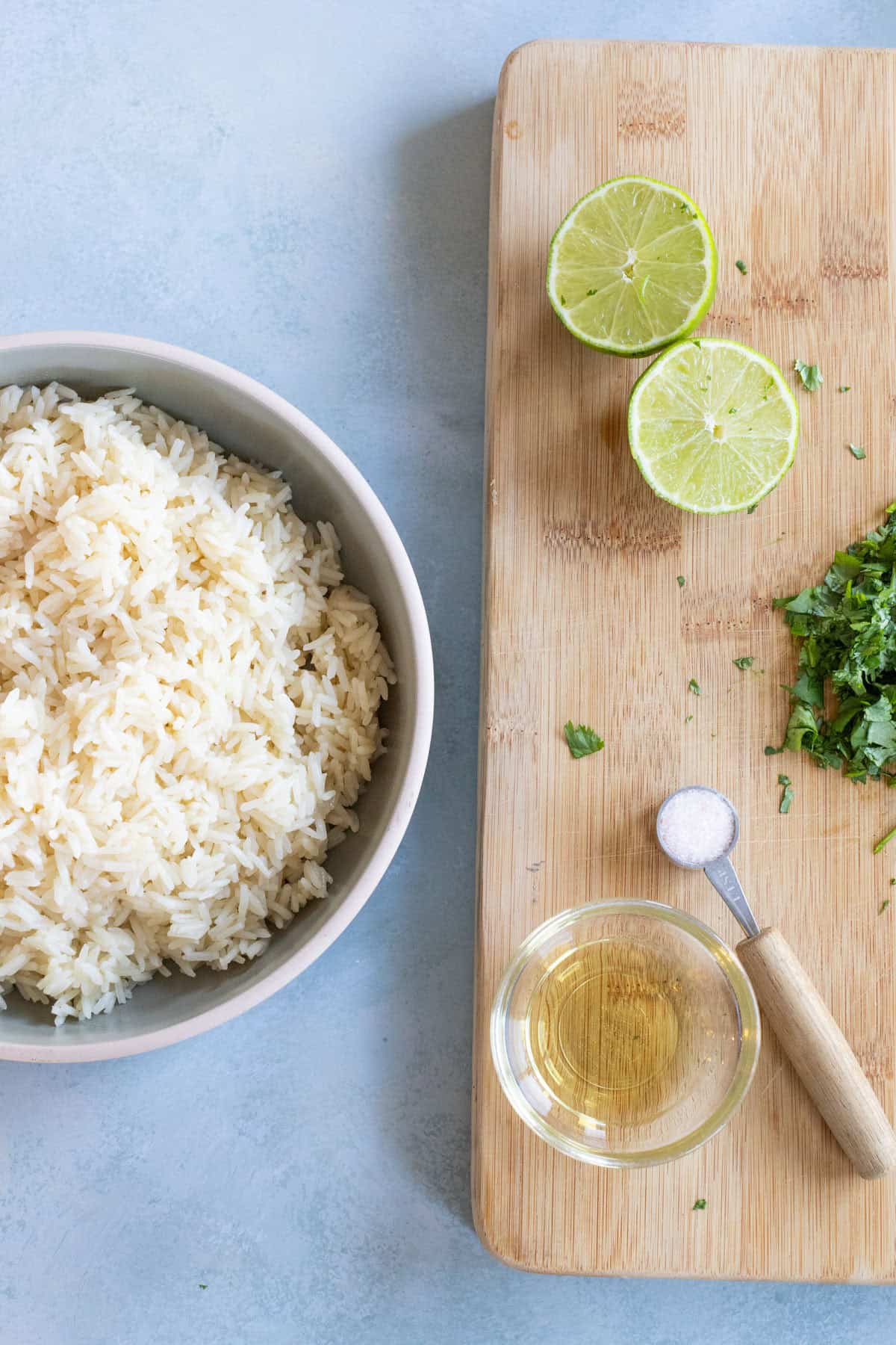 A bowl of cooked white rice next to a cutting board with halved limes, chopped herbs, a small glass of oil, and a measuring spoon with salt.
