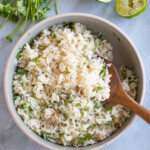 A wooden spoon serving cilantro lime rice from a sauce pot that sits on a light gray background. Fresh cilantro sprigs and lime halves are nearby.