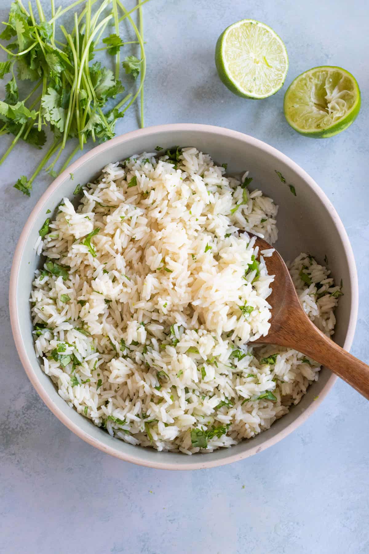 A wooden spoon serving cilantro lime rice from a sauce pot that sits on a light gray background. Fresh cilantro sprigs and lime halves are nearby.