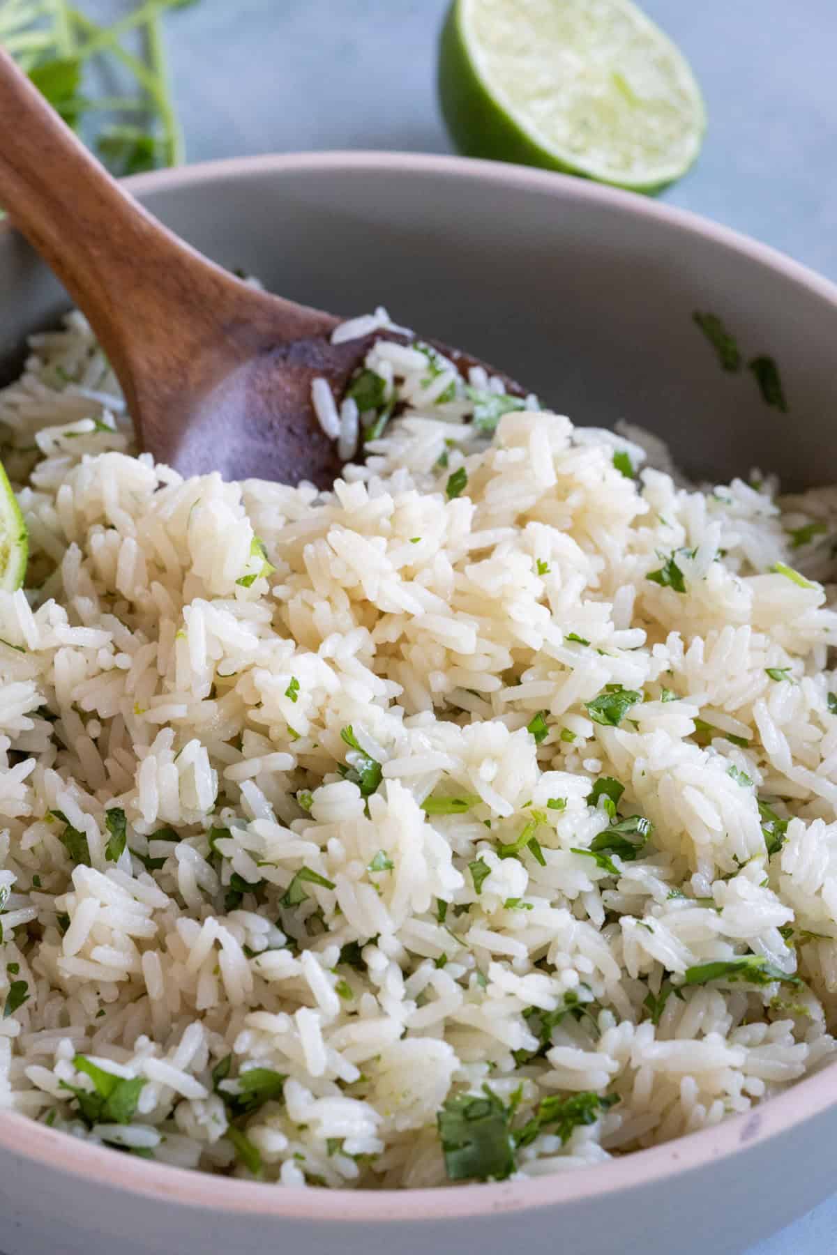 A bowl of white rice mixed with chopped cilantro, with a wooden spoon and a halved lime in the background.