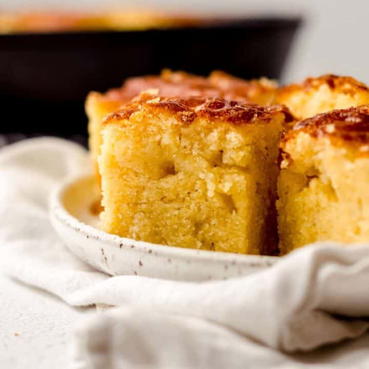 Close-up of three pieces of cornbread on a white plate, with a cloth napkin underneath and a blurred pan in the background.
