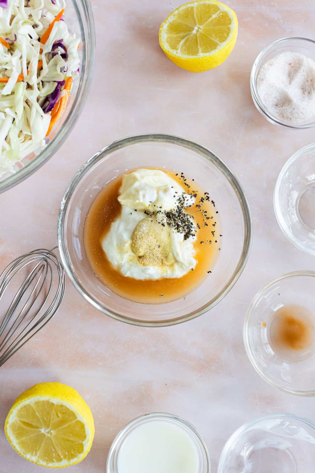 A glass bowl with mayonnaise, mustard, spices, and liquid ingredients sits on a counter surrounded by lemon halves, a whisk, and other small bowls of ingredients.